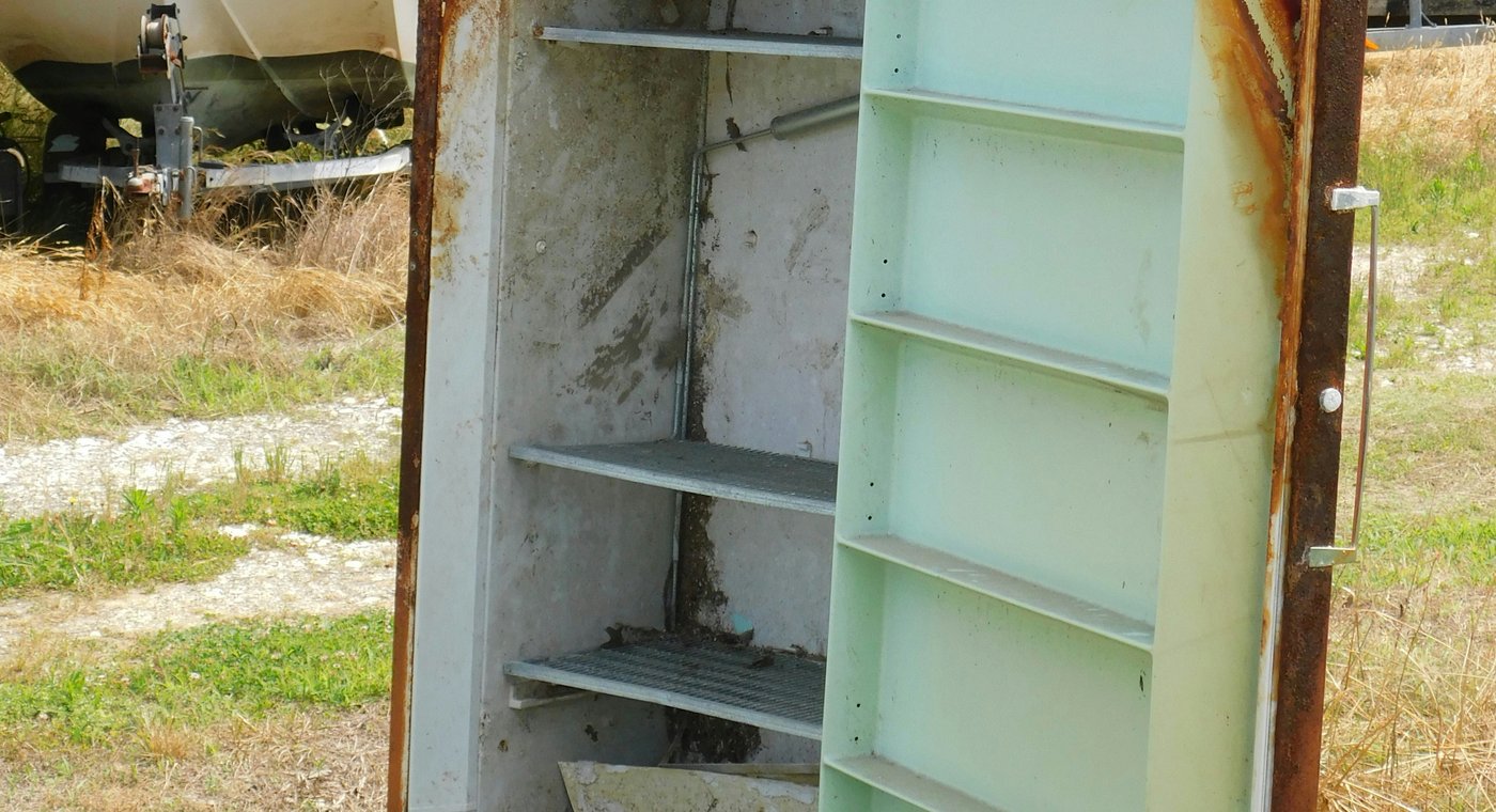 An old rusted refrigerator standing open in dry grass, its mint-green door shelves showing