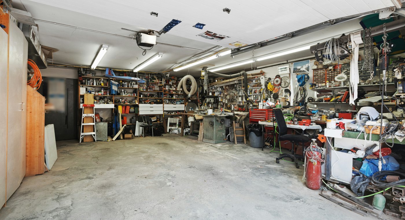 A deep two-car garage crammed with shelving, tools, a table saw, a red tool chest and hoses over a bare concrete floor