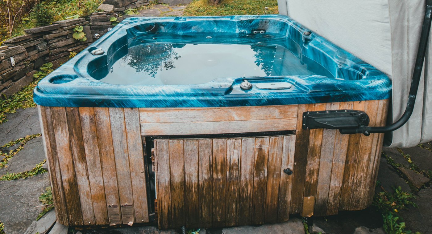 An old acrylic hot tub with a marbled blue shell and a weathered cedar skirt, its cover folded back, on flagstone