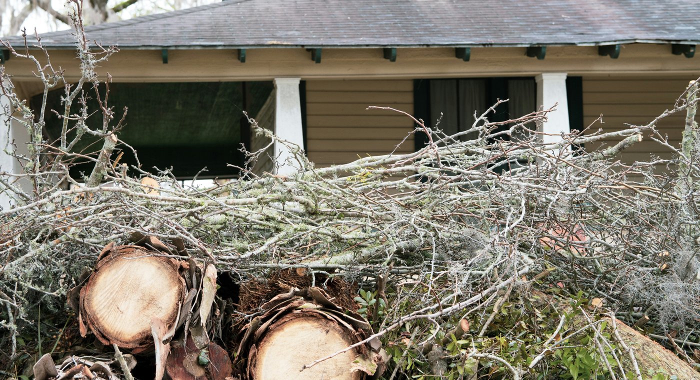 A pile of cut branches and log rounds heaped in front of the porch of a tan clapboard house